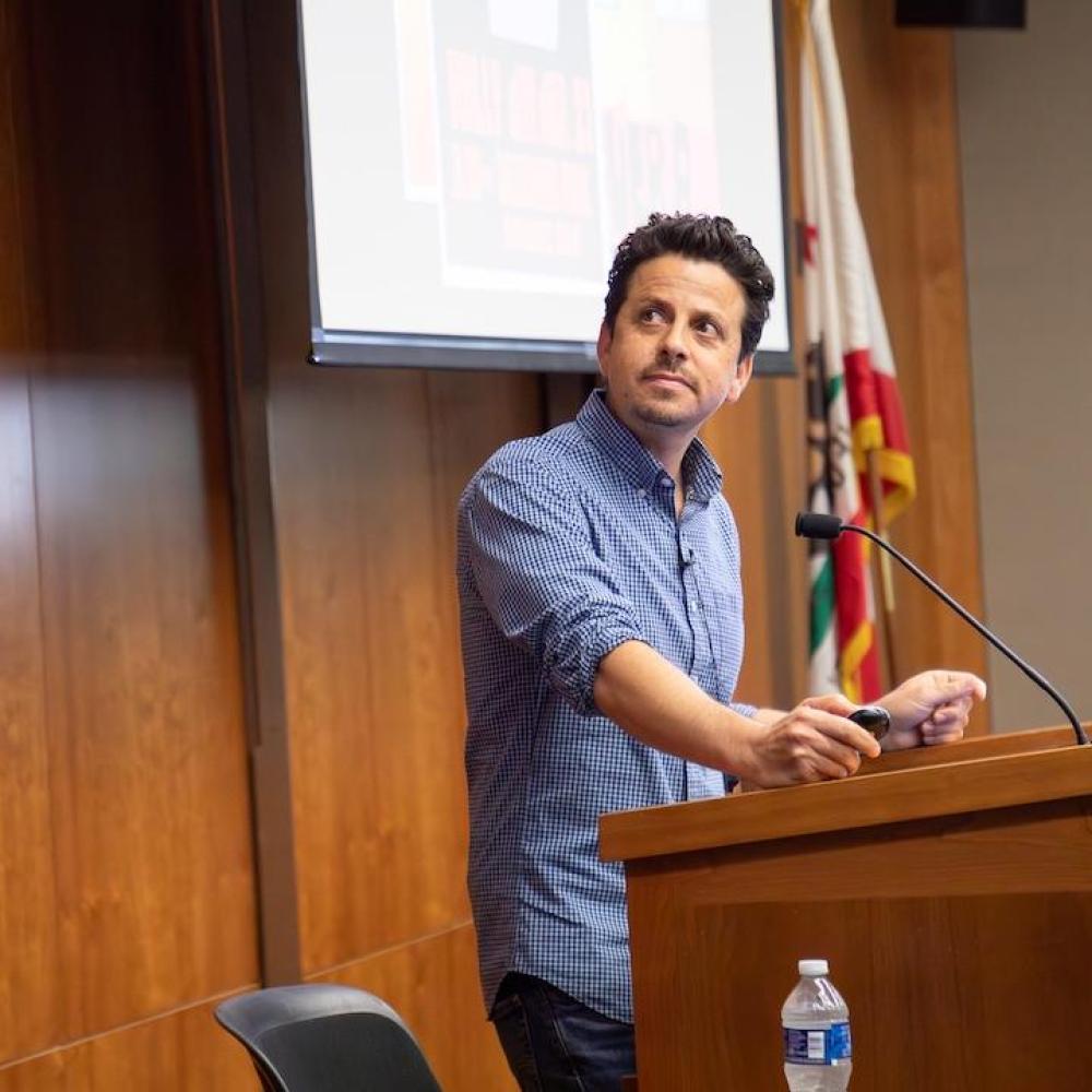 Focused man in blue shirt speaking at wooden podium in lecture hall, projection screen behind him