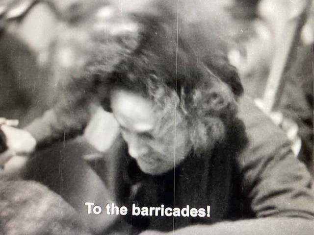 Black-and-white closeup still from Les Mis of a person struggling. Subtitle reads, "To the barricades!"