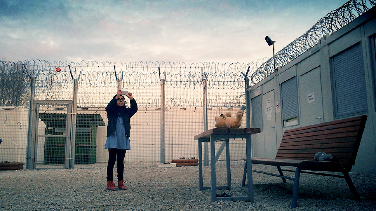 A still from Midnight Traveler Still from Midnight Traveler: a child stands in an enclosure surrounded by barbed wire, taking a video with a cell phone.