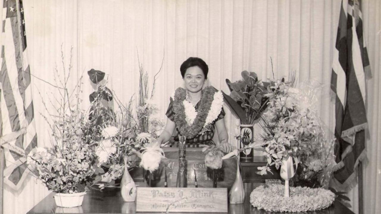 Wu described how when Patsy Mink left Hawaii for Washington, D.C., her constituents "sent the Pacific with her" in the form of leis and plants. Black and white photo of Patsy Mink standing behind her desk, surrounded by plants and wearing several leis