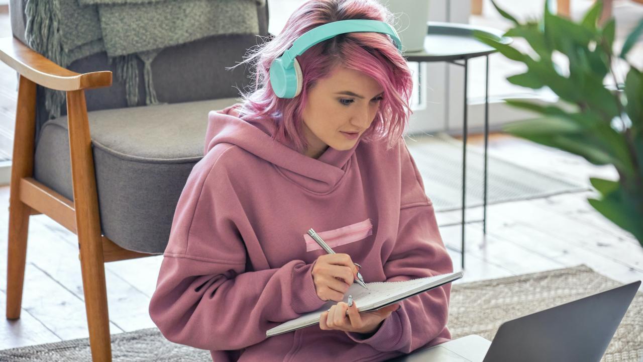 Image of person with pink hear wearing headphones writing in a notebook while looking at a laptop