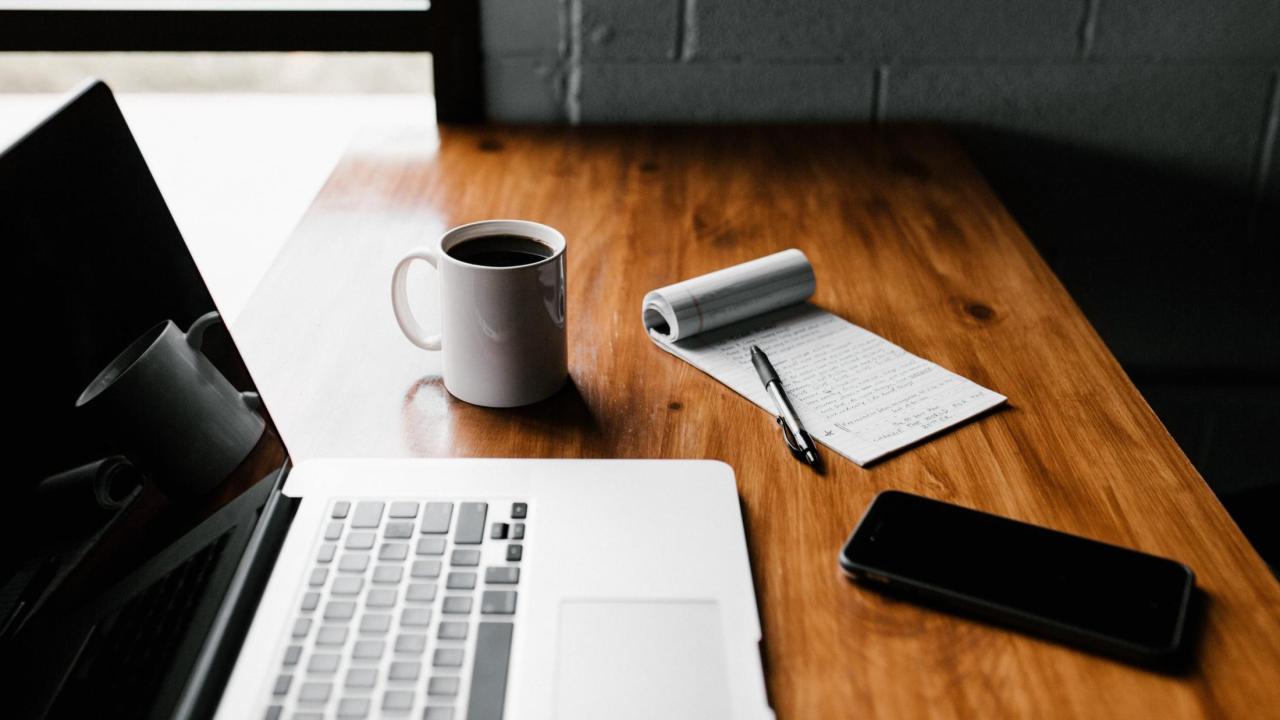 A laptop, coffee mug, notepad, and iPhone on a table