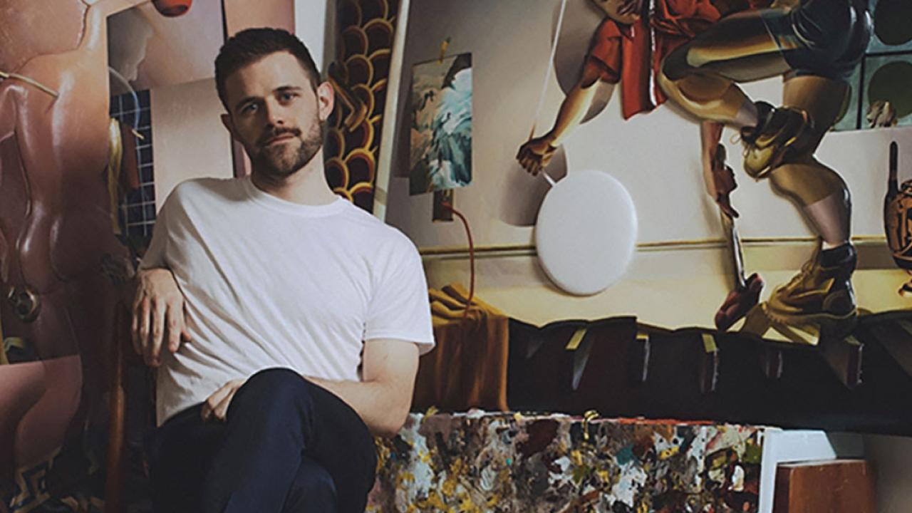 Kyle Dunn wears brown boots and a white tee and sits in a chair next to an abstract painting