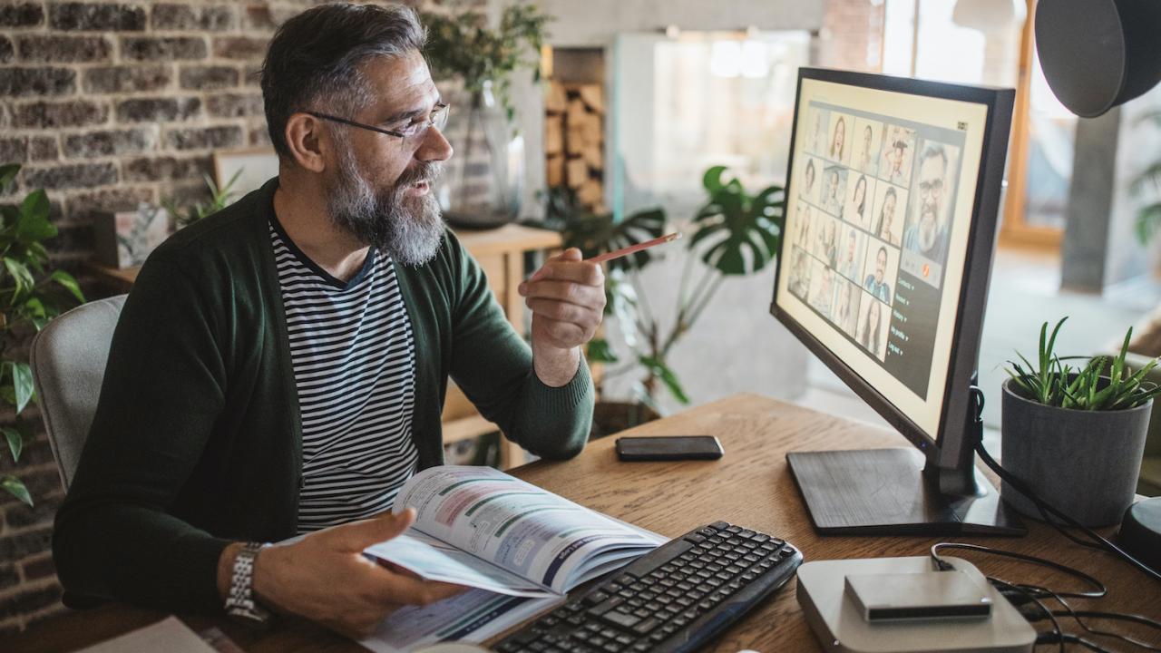 Person with a beard sits in front of a desktop computer, gesturing toward faces in a Zoom call on the screen