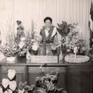 Black and white photo of Patsy Mink standing behind her desk, surrounded by plants and wearing several leis