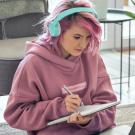 Image of person with pink hear wearing headphones writing in a notebook while looking at a laptop